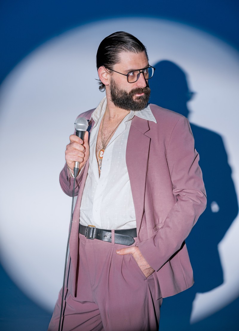 [small] Portrait of Ben Gorodetsky in a sharp pink suit, open dress shirt and holding a microphone. Photo credit Bangishimo